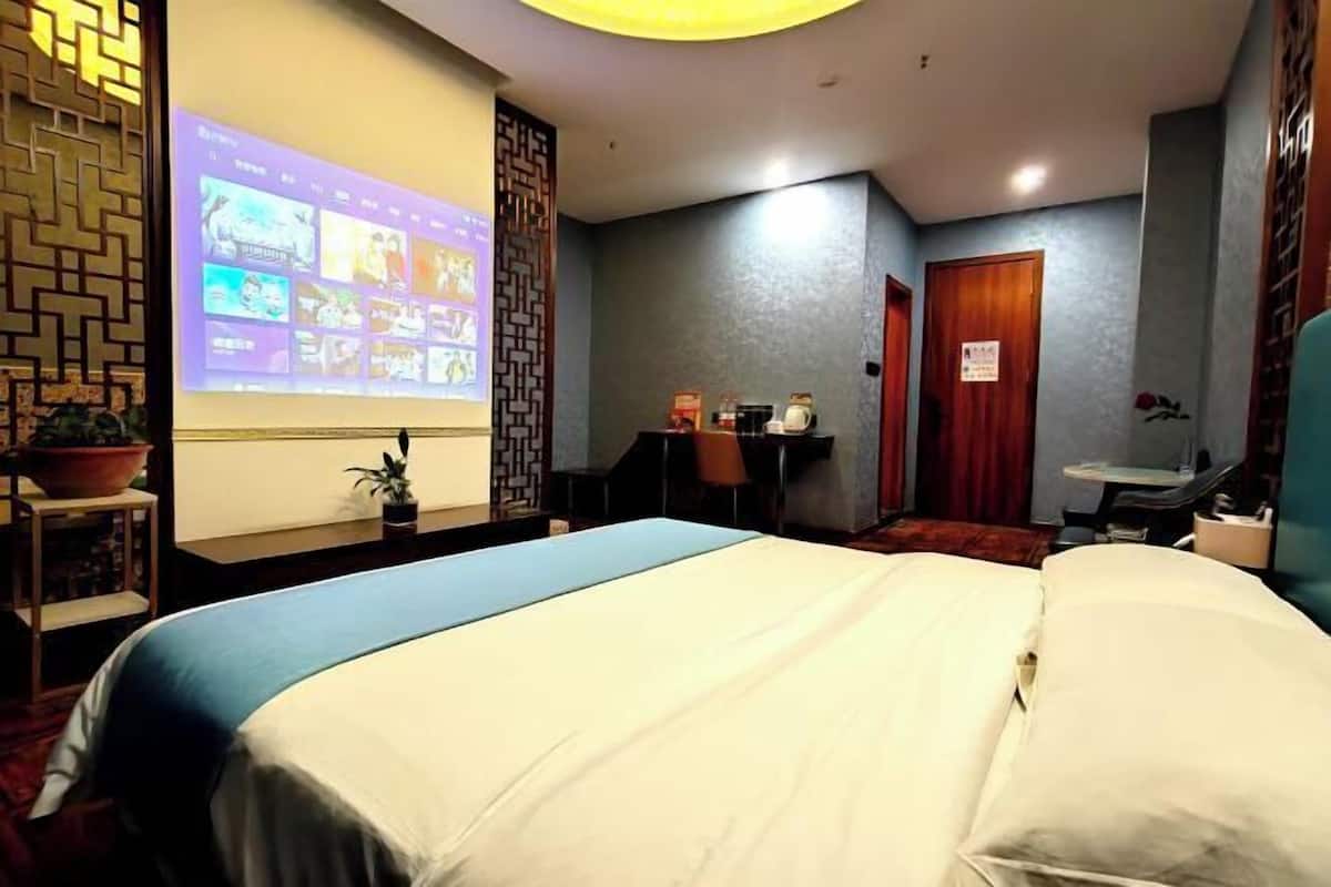 Hotellihuone, jossa on sänky, työpöytä tuolilla, pieni pöytä, jossa on kasvi, ja projektorikangas, jolle heijastetaan kuvia.