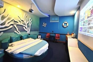 Habitación de hotel con cama, escritorio, dos sillas, pared con motivos náuticos y una mesita de noche con lámpara.