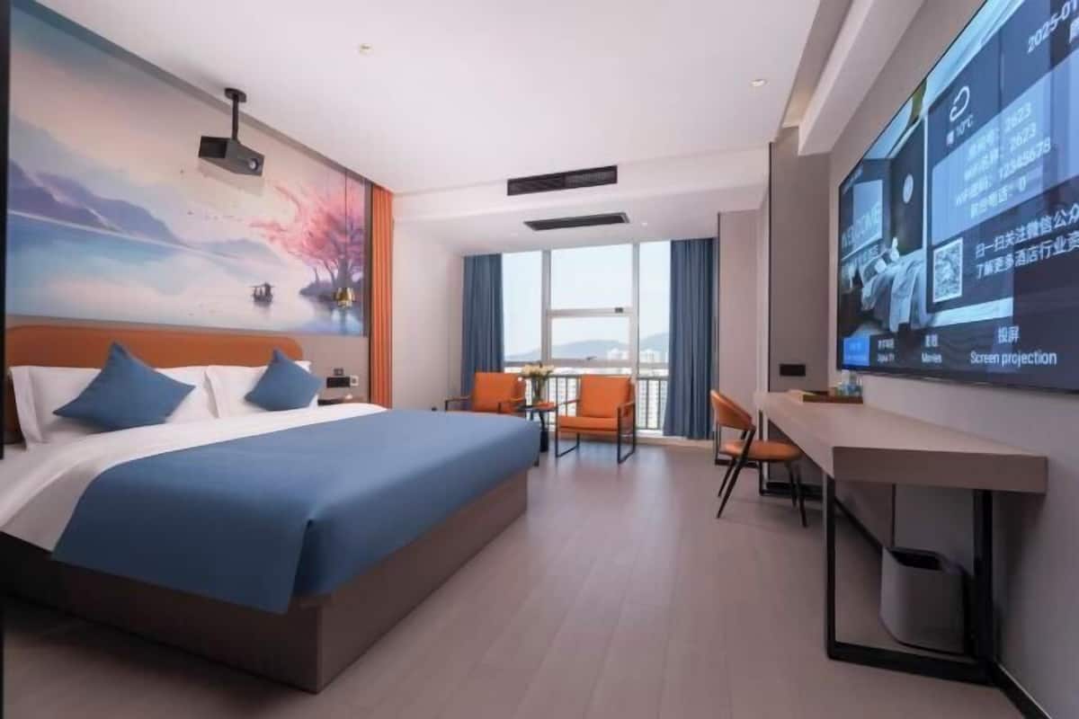 A modern hotel room with a large bed, a desk, a chair, a TV screen, and a balcony with a view.
