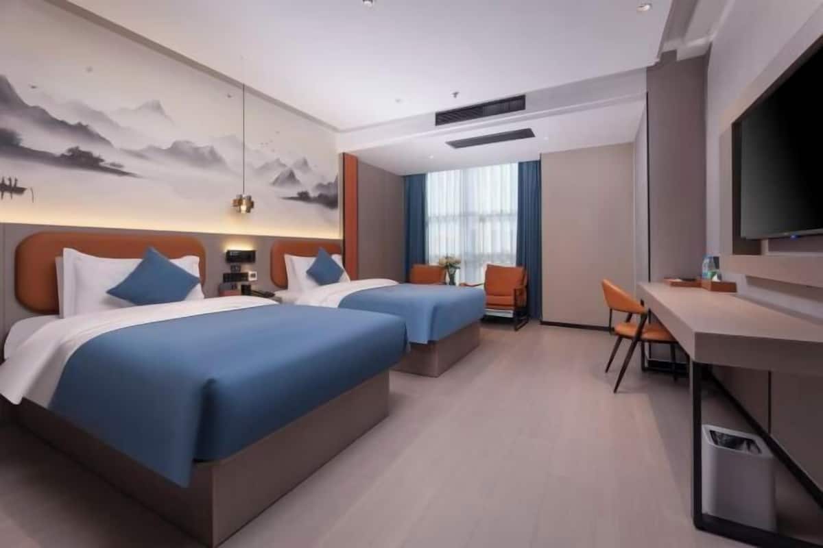 A hotel room with two beds, a desk, a chair, a TV, and a wall with a mountain painting.