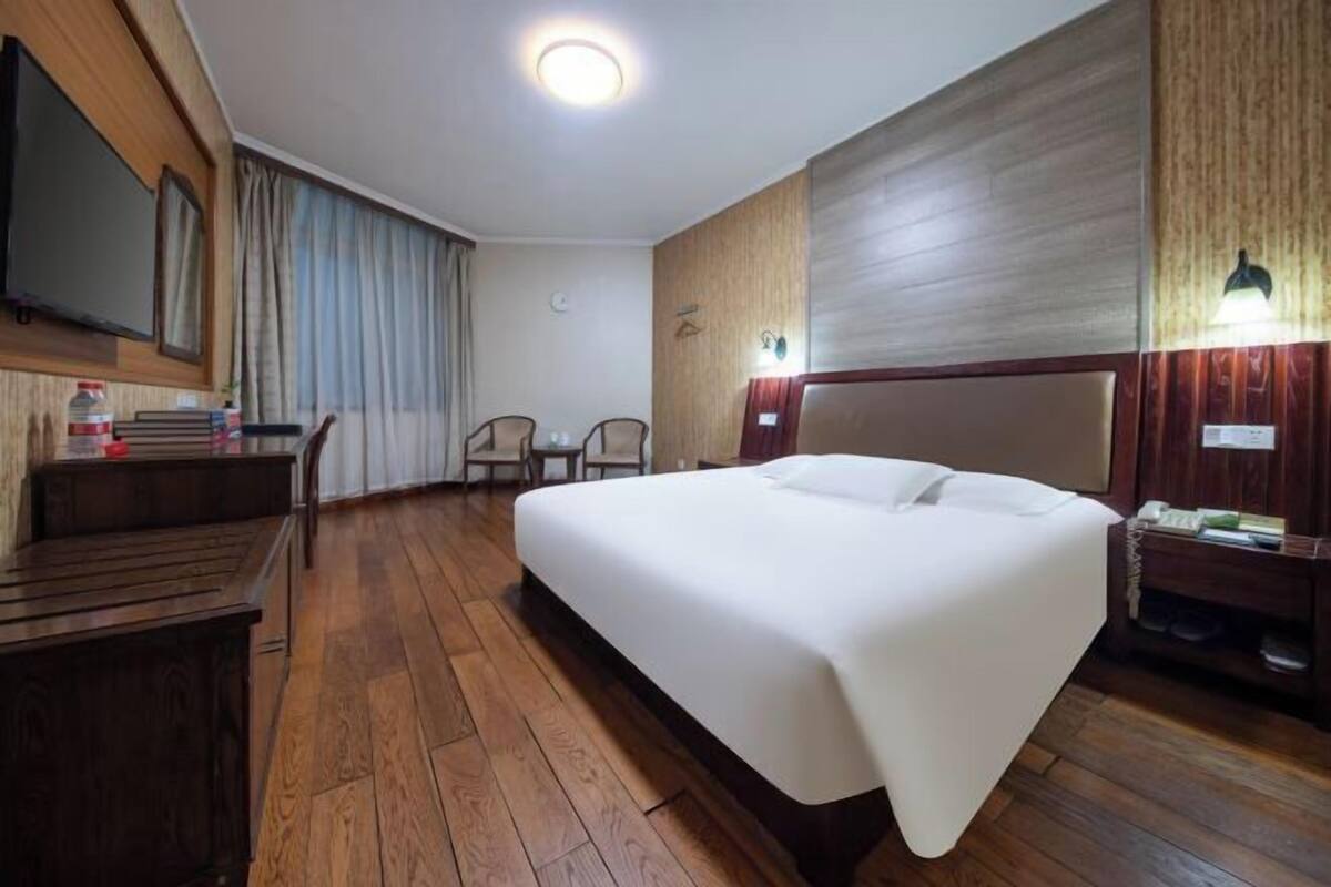 Habitación de hotel con una cama grande, suelo de madera, un escritorio con silla, un televisor y una mesita de noche con lámpara.