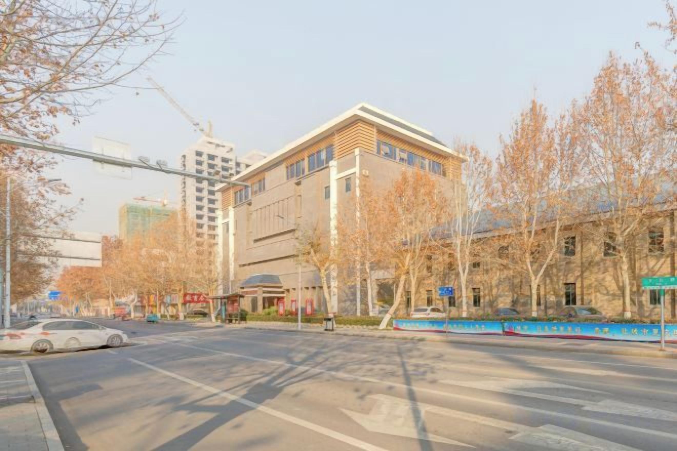 A street view with a building that has multiple floors and a flat roof. There are trees with no leaves, suggesting it might be autumn or winter.