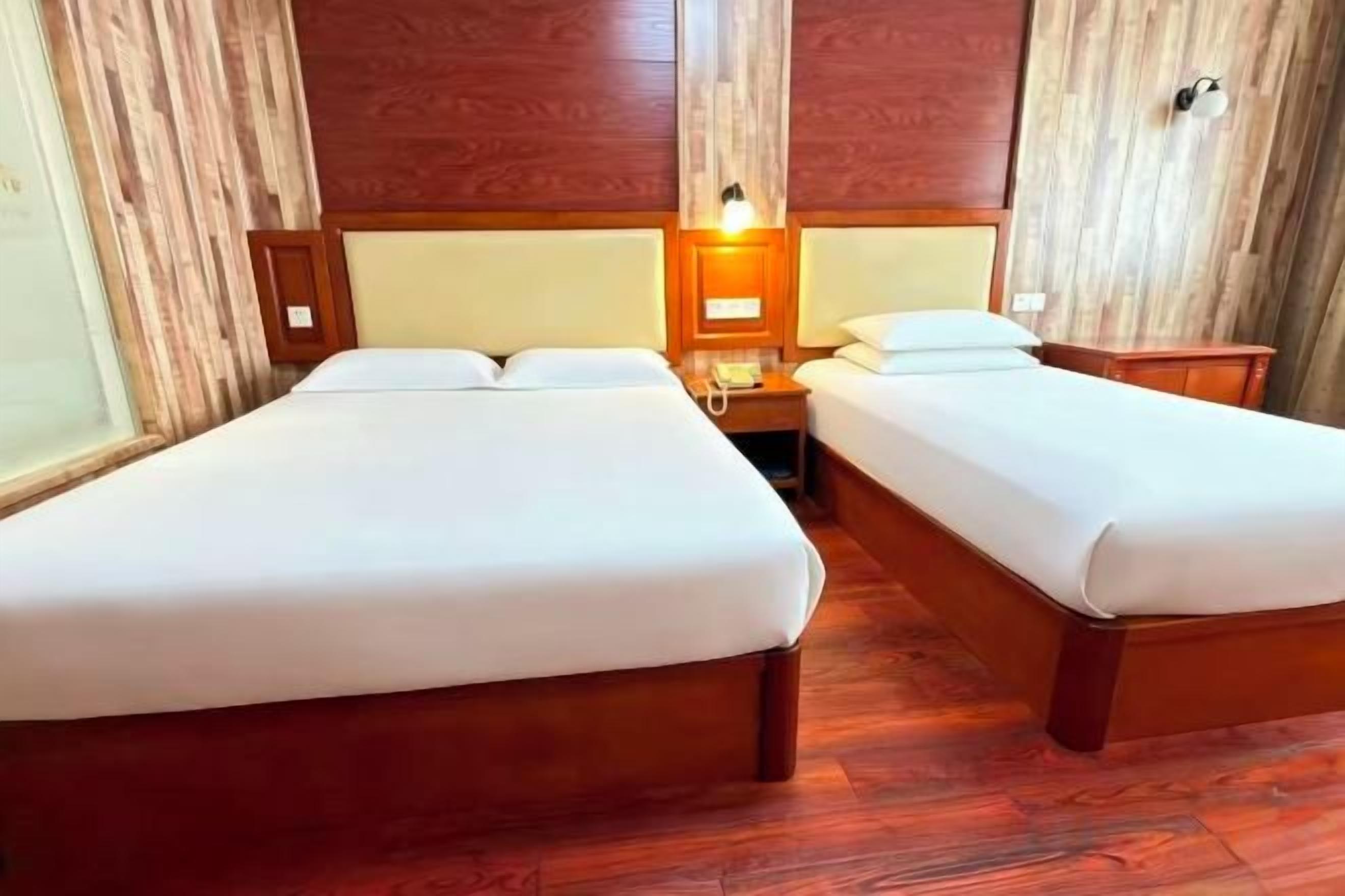 A hotel room with two single beds, wooden headboards, a nightstand with a lamp, and a wooden floor.