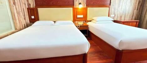 A hotel room with two single beds, wooden headboards, a nightstand with a lamp, and a wooden floor.