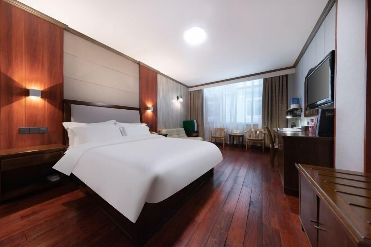 Ein Hotelzimmer mit einem großen Bett, Holzboden, einem Schreibtisch mit Stuhl, einem Fernseher und einem Fenster mit Vorhängen.