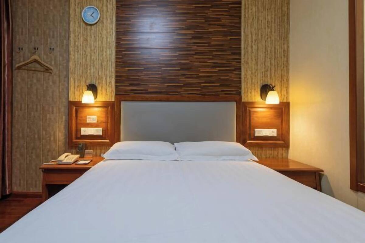 Habitación de hotel con cama, cabecera de madera, lámparas de noche, un reloj y un teléfono en la mesita.