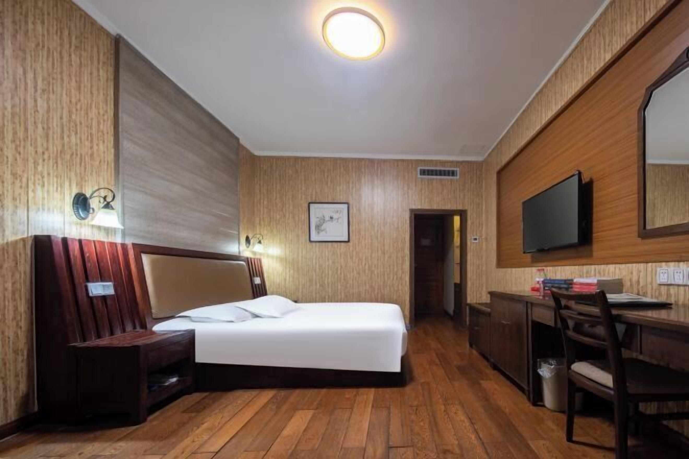 A hotel room with a bed, bedside table, lamp, TV, desk, chair, and a mirror on the wall.