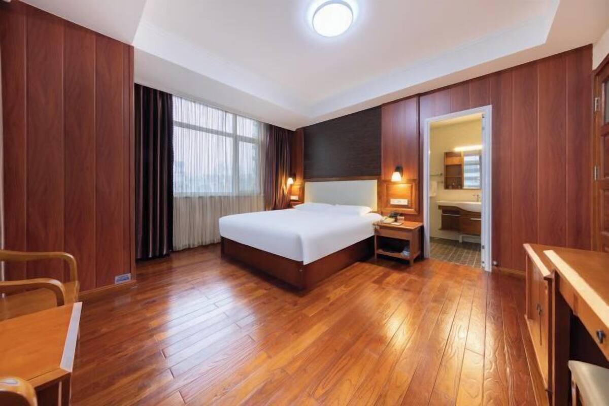 Habitación de hotel con piso de madera, cama, escritorio, silla, mesita de noche, lámpara, ventana con cortinas y puerta que da a un baño.