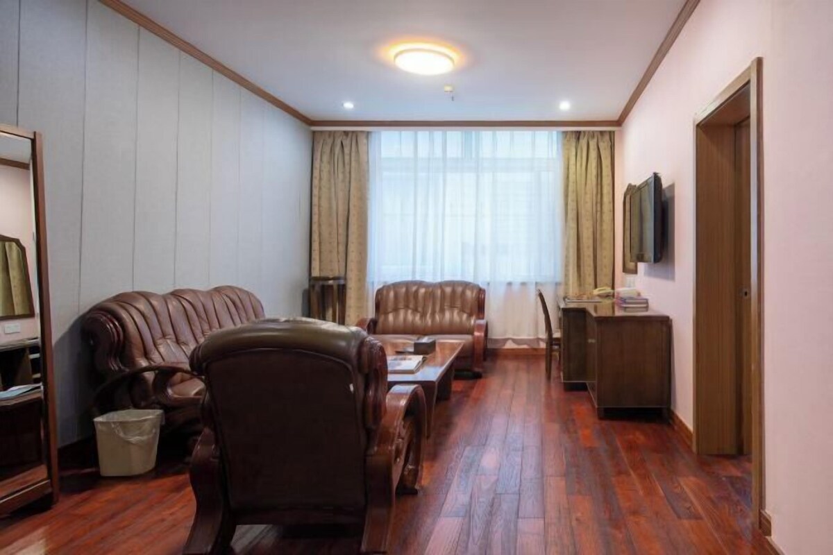Ein Zimmer mit Ledersofa, einem Holztisch, einem Schreibtisch mit Stuhl, einem Fernseher und einem Fenster mit Vorhängen.