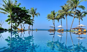 A clear blue pool with reflections of palm trees and lounge chairs, surrounded by a tropical setting with a view of the ocean. - One Myanmar Resort Ngwe Saung (Ngwe Saung)