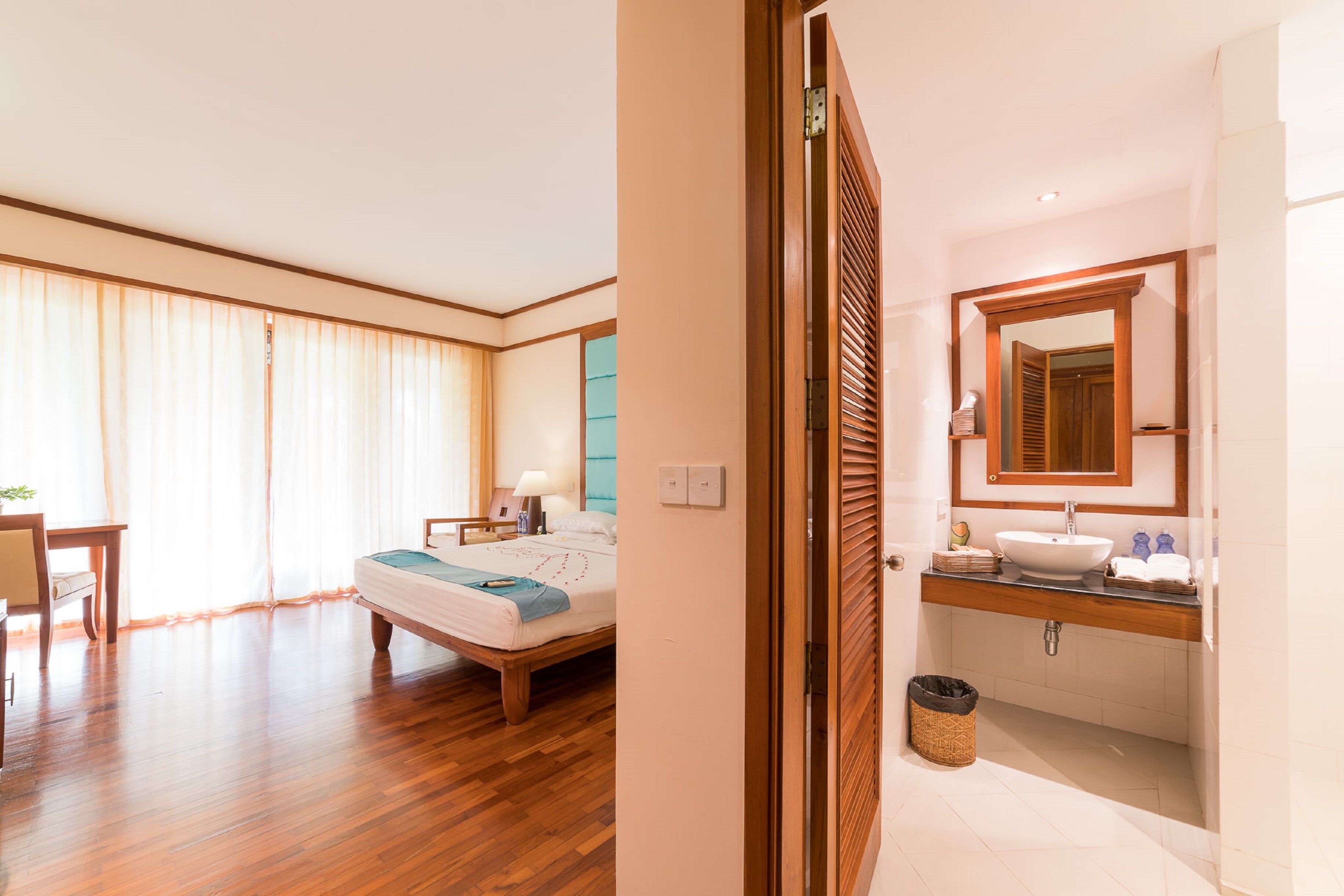 Habitación de hotel con piso de madera, cama con manta azul, mesita de noche con lámpara, y baño con tocador de madera y un espejo grande.