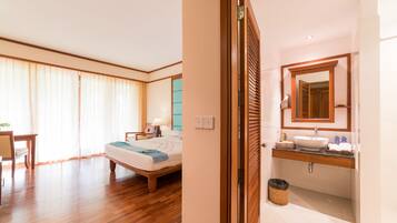 Habitación de hotel con piso de madera, cama con manta azul, mesita de noche con lámpara, y baño con tocador de madera y un espejo grande.