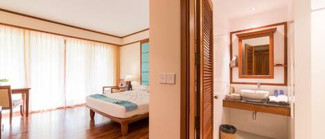 A hotel room with a wooden floor, a bed with a blue blanket, a bedside table with a lamp, a bathroom with a wooden vanity and a large mirror.
