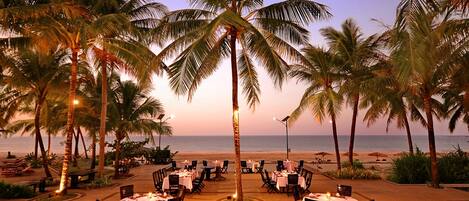 Un área de comedor frente a la playa con mesas y sillas dispuestas alrededor de una palmera, con vista al océano.