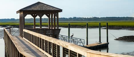 A wooden pier leading to a gazebo by a calm body of water, surrounded by grassy fields.
