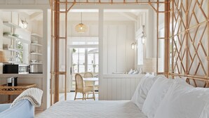 A bedroom with a large bed, a wicker chair, a dining table, and a kitchen area visible through an open door. - "The Goldeneye” (3021 Southerly Way) 2 person creekside bungalow at Kiawah River (Johns Island)