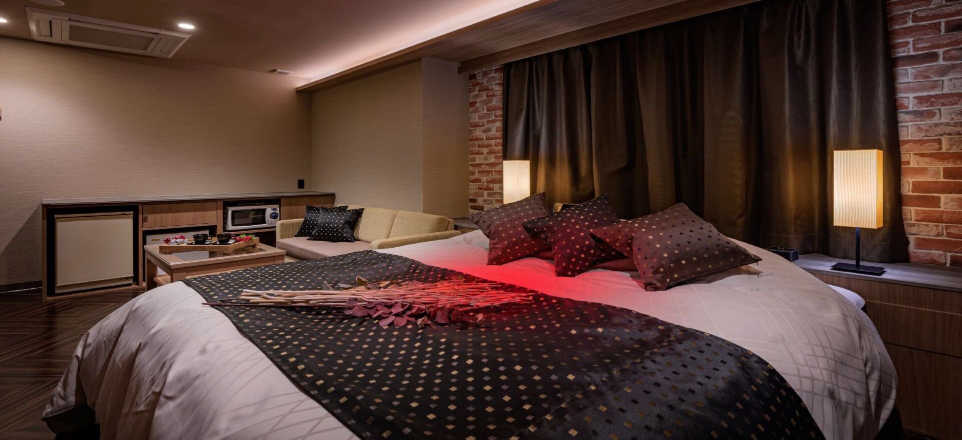 A hotel room with a large bed, a desk with a computer, a small kitchenette, and a brick wall.