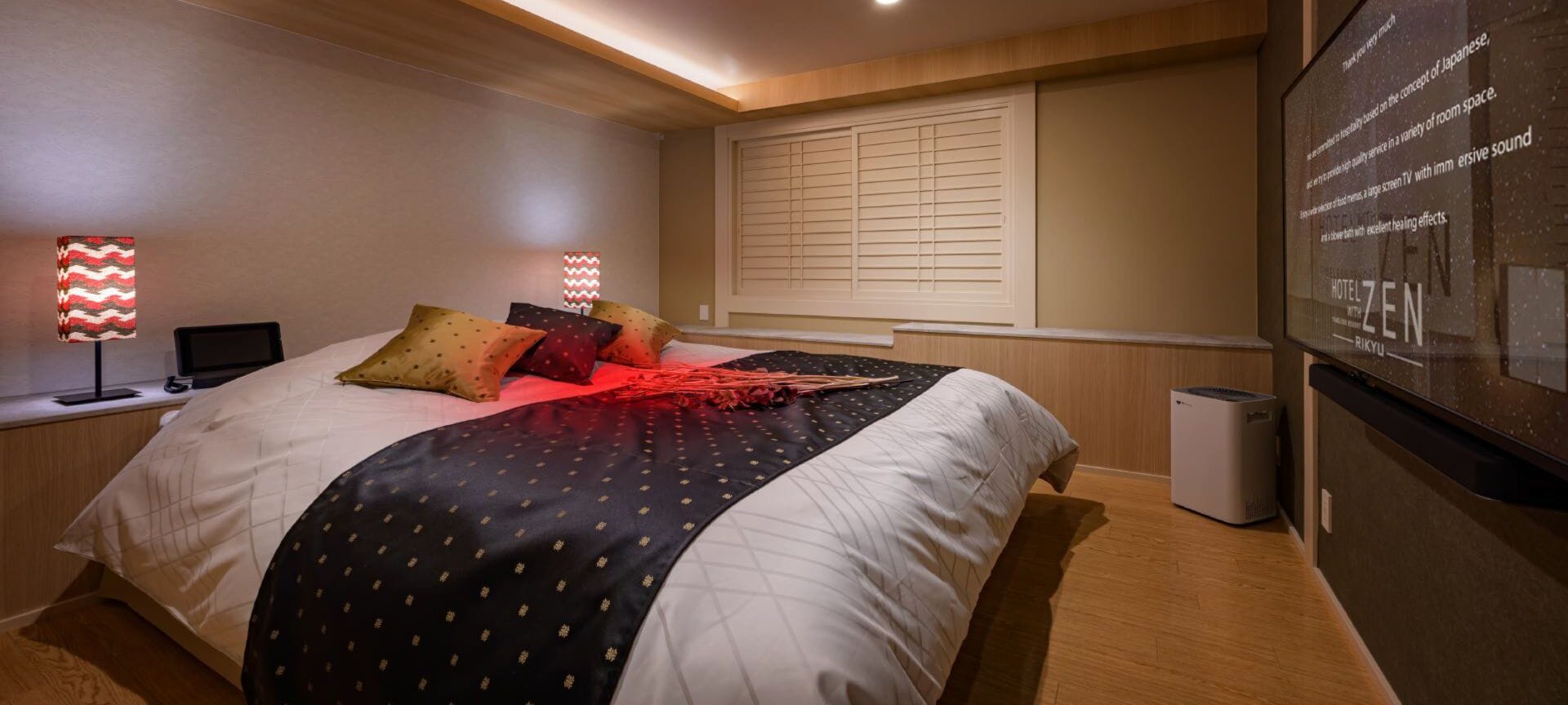 Una camera d'albergo con un letto, due lampade sui comodini, un'ampia finestra con persiane, una TV a parete e un cestino.