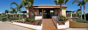 A building with a covered entrance, a staircase leading up to it, and a landscaped area with palm trees. - Residence Hydria (Campofelice di Roccella)