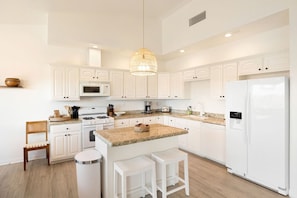 A modern kitchen with white cabinetry, granite countertops, a central island with stools, a built-in oven, and a refrigerator. - Rancho Nopales JT-Pool, Hot Tub, Hammocks & Views (Joshua Tree)
