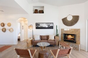 A living room with a sofa, a glass coffee table, and two wicker chairs. There is a fireplace with a fire, a framed picture on the wall, and a hanging basket. - Rancho Nopales JT-Pool, Hot Tub, Hammocks & Views (Joshua Tree)