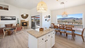 A spacious living area with a fireplace, dining table, and chairs, a kitchen with an island, and a large window with a mountain view. - Rancho Nopales JT-Pool, Hot Tub, Hammocks & Views (Joshua Tree)