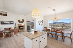 A spacious living area with a fireplace, dining table, and chairs, a kitchen with an island, and a large window with a mountain view. - Rancho Nopales JT-Pool, Hot Tub, Hammocks & Views (Joshua Tree)