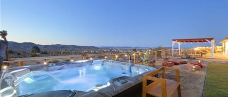 A hot tub with a wooden deck and a pergola with string lights, overlooking a cityscape at dusk.