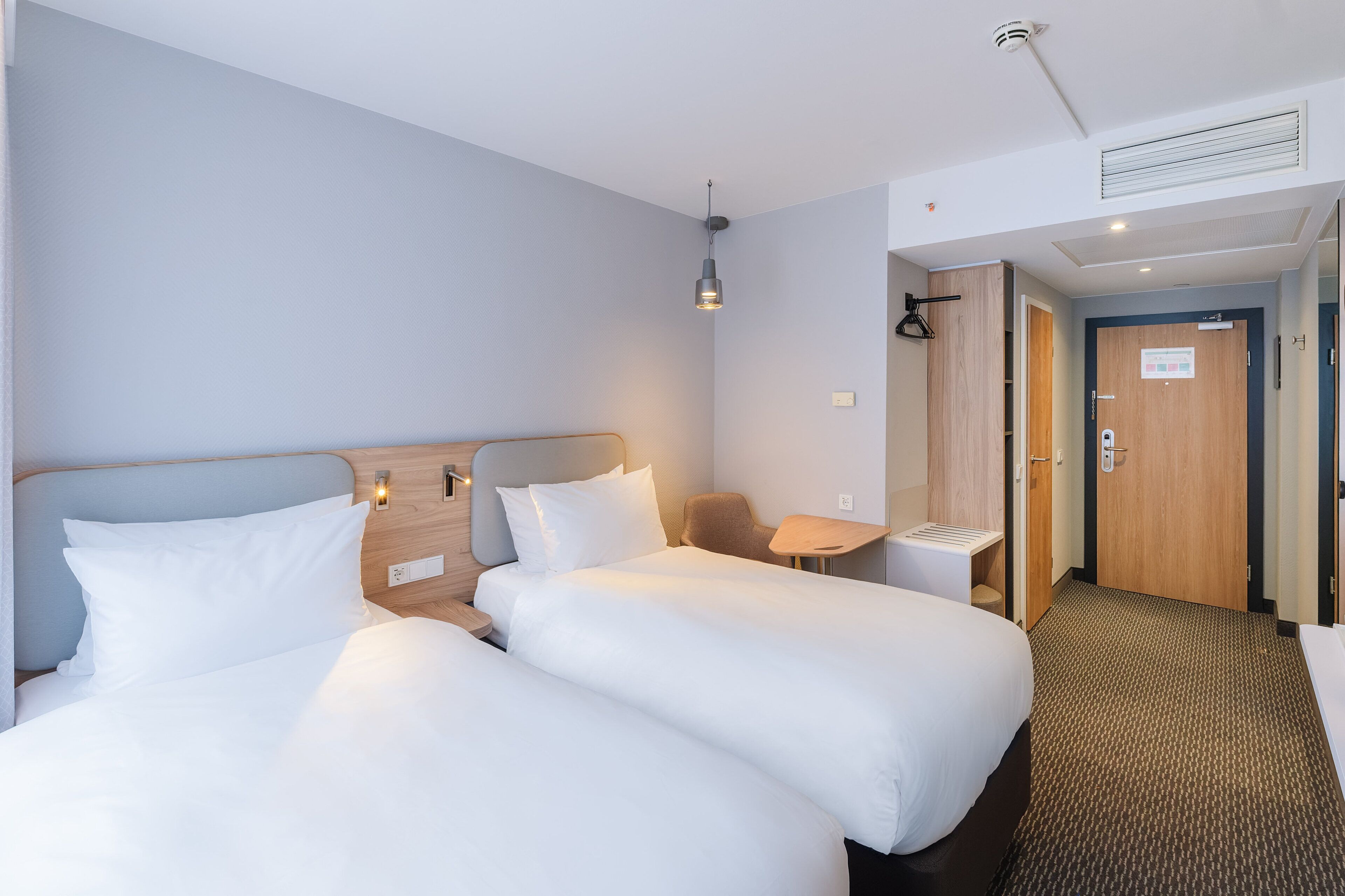 A hotel room with two beds, a small table, a chair, and a door leading to a bathroom.