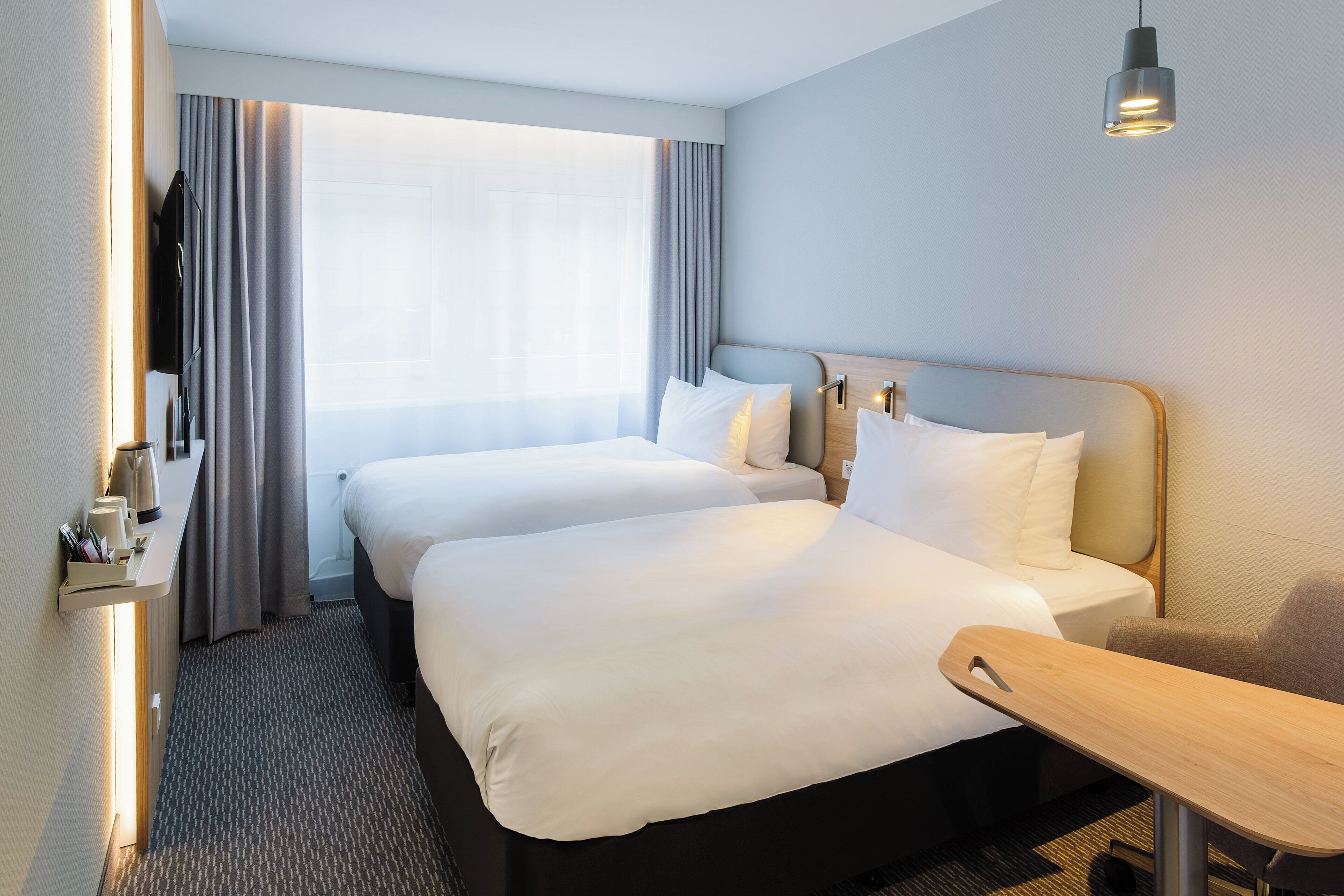 Ein Hotelzimmer mit zwei Betten, einem Schreibtisch, einem Stuhl, einer Kaffeemaschine und einem Fenster mit Vorhängen.