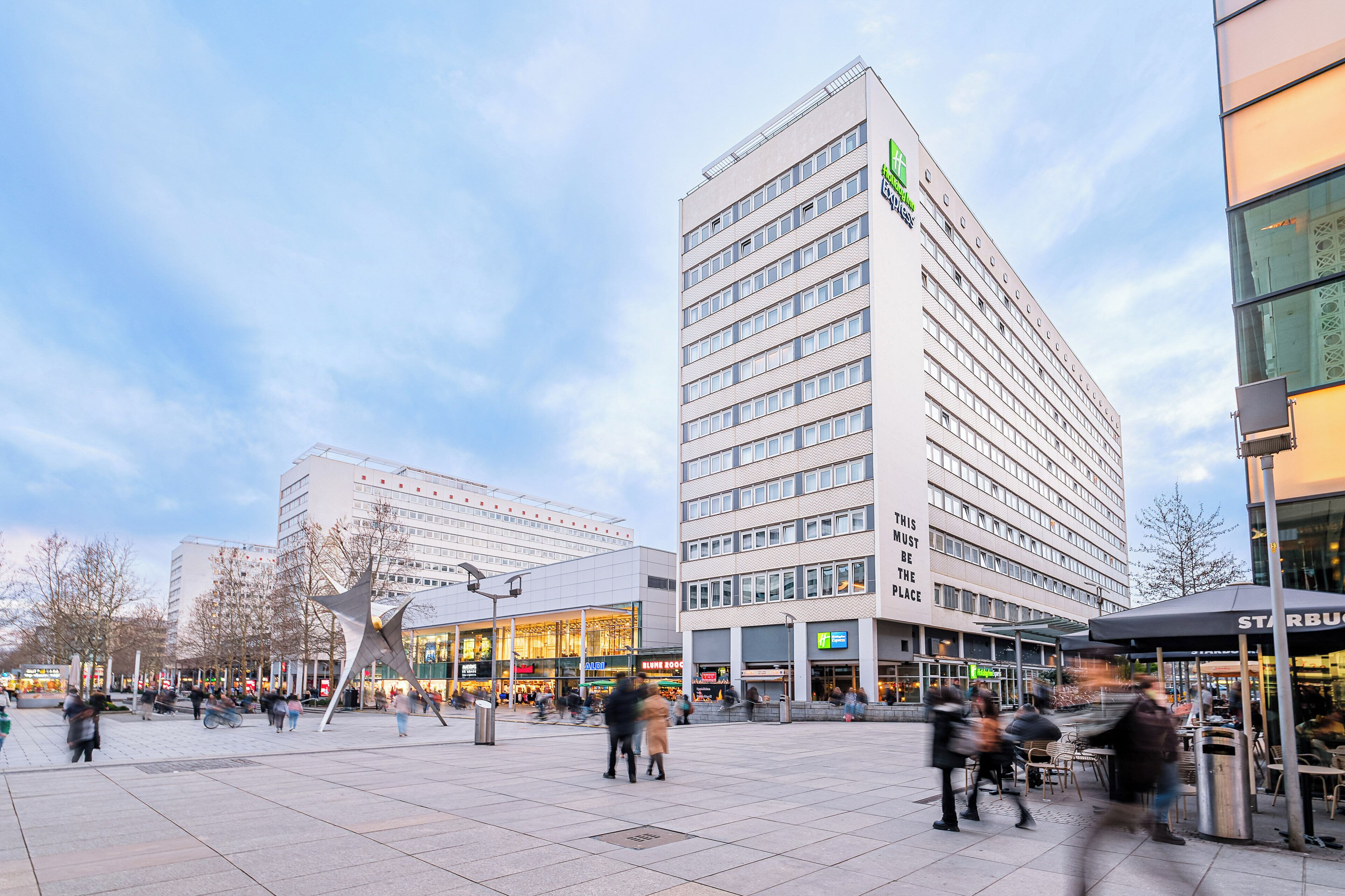 A modern hotel with a prominent green logo, a spacious plaza with seating areas, and a Starbucks café.
