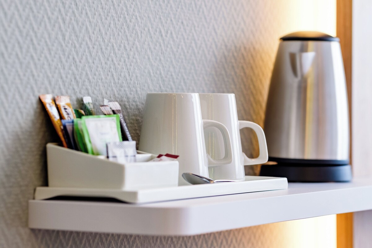 Una taza blanca sobre una estantería, con una bandeja blanca que contiene varios objetos, incluyendo un paquete verde, un paquete rojo y un paquete blanco.
