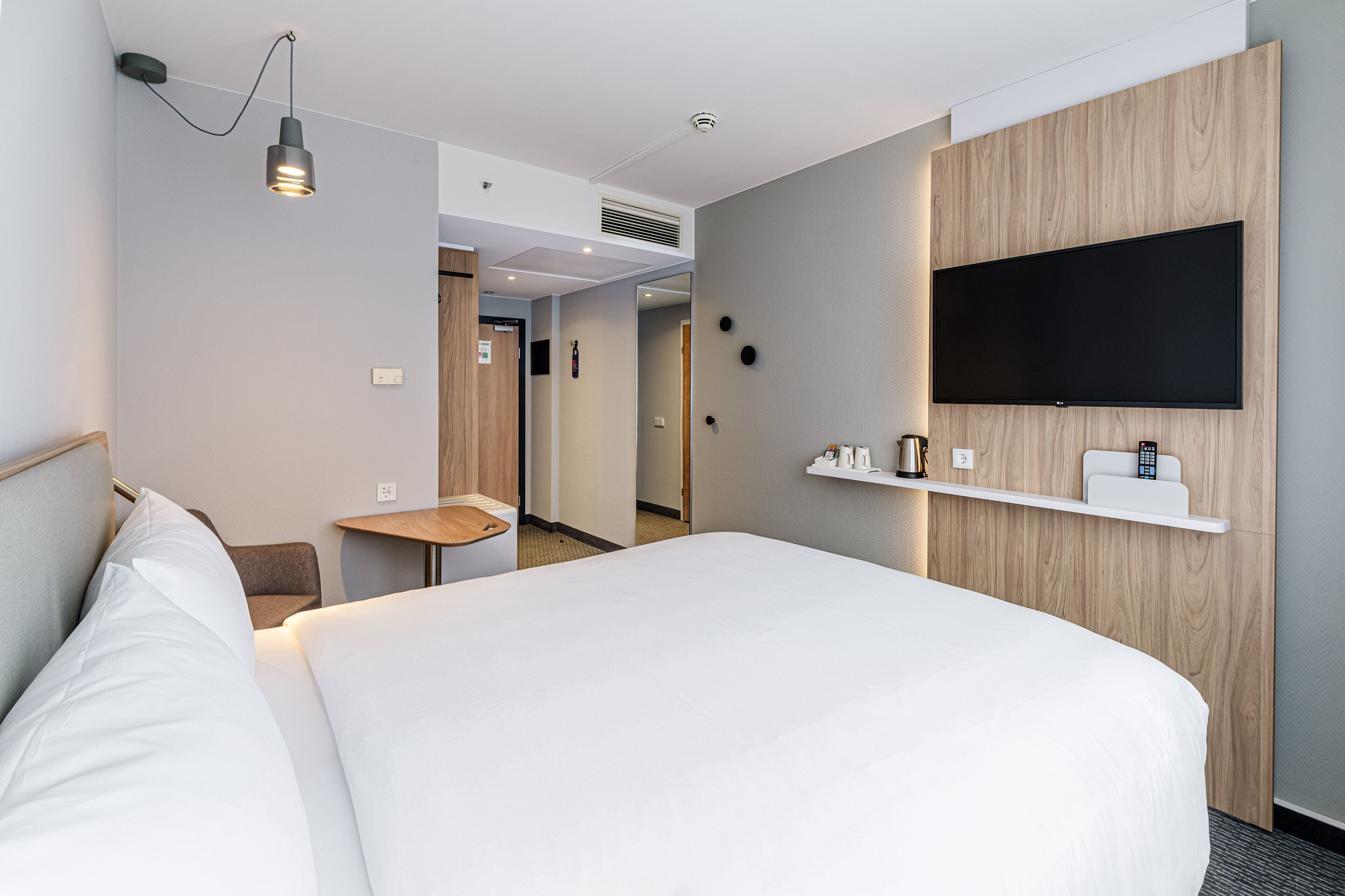 A hotel room with a bed, bedside table, a mounted TV, and a small desk area.