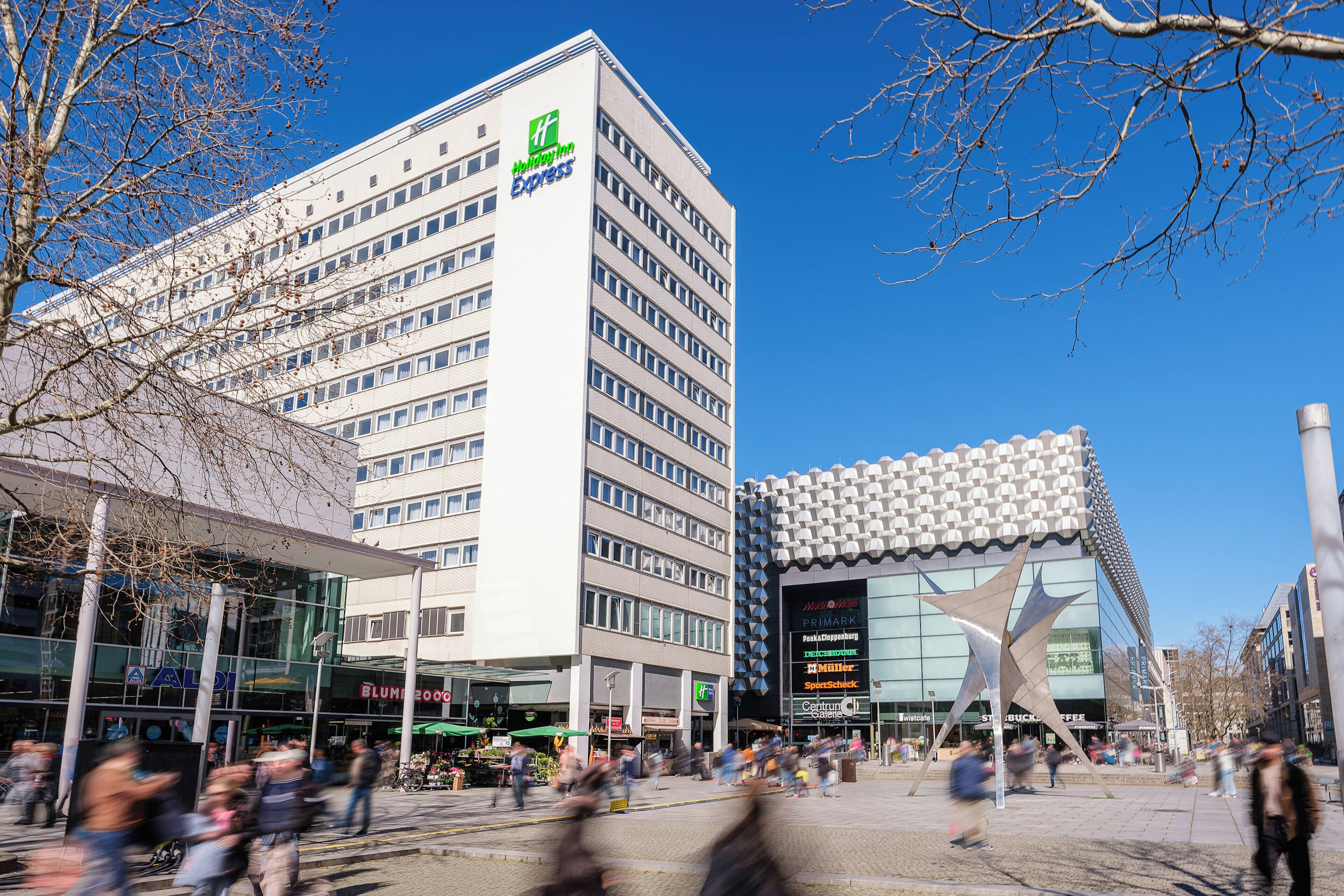 A modern hotel with a prominent green logo, a glass facade, and a distinctive star-shaped sculpture in front.