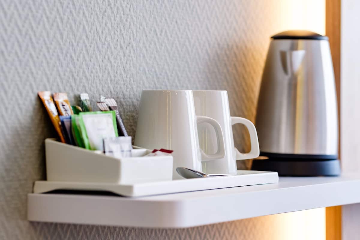 Une tasse blanche est posée sur une étagère blanche, à côté d’une bouilloire argentée et d’un petit plateau blanc contenant des stylos et un carnet.