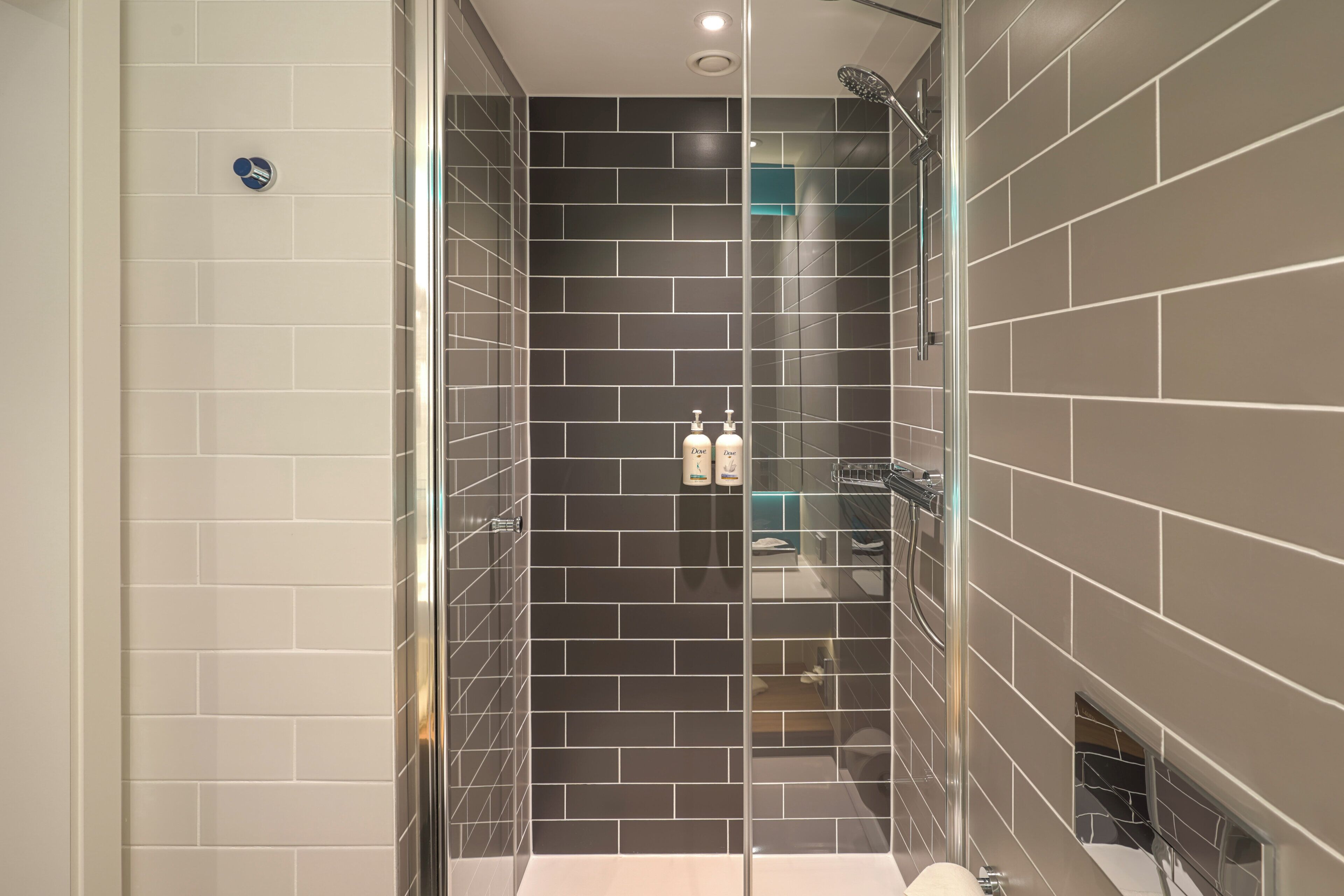 Un baño moderno con ducha envuelta en vidrio, paredes de azulejos oscuros y un lavabo blanco.