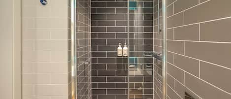 Un baño moderno con ducha envuelta en vidrio, paredes de azulejos oscuros y un lavabo blanco.