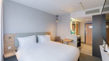 Habitación de hotel moderna con una cama grande, mesitas de noche, una pequeña mesa redonda, una silla, un espejo y un baño visible a través de una puerta abierta.