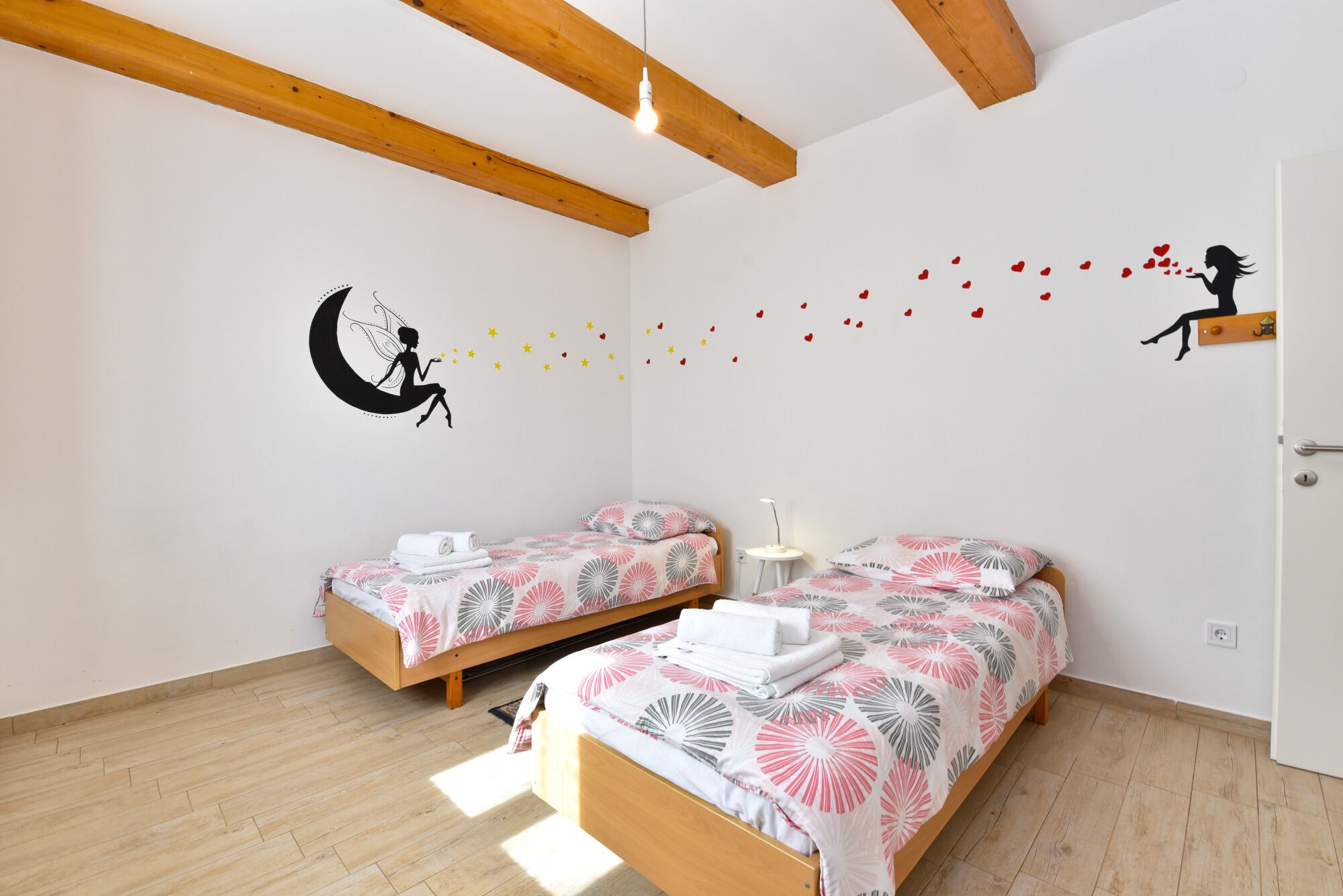 A bedroom with two beds, wooden beams, a wall decal of a woman on a crescent moon, and a door with a small shelf.