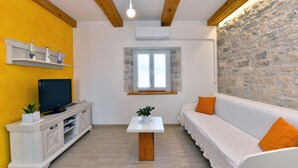 A room with a stone wall, a white sofa with orange cushions, a white TV stand, and a small table with a plant. - House 2176/55604 (Istria - Debeljuhi), 19000m from the beach (Debeljuhi)