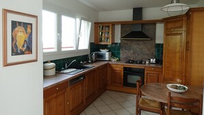 Ett kök med träskåp, en mikrovågsugn, en spis och ett diskho. Det finns ett fönster med gardiner, en inramad tavla på väggen och ett matbord med stolar.