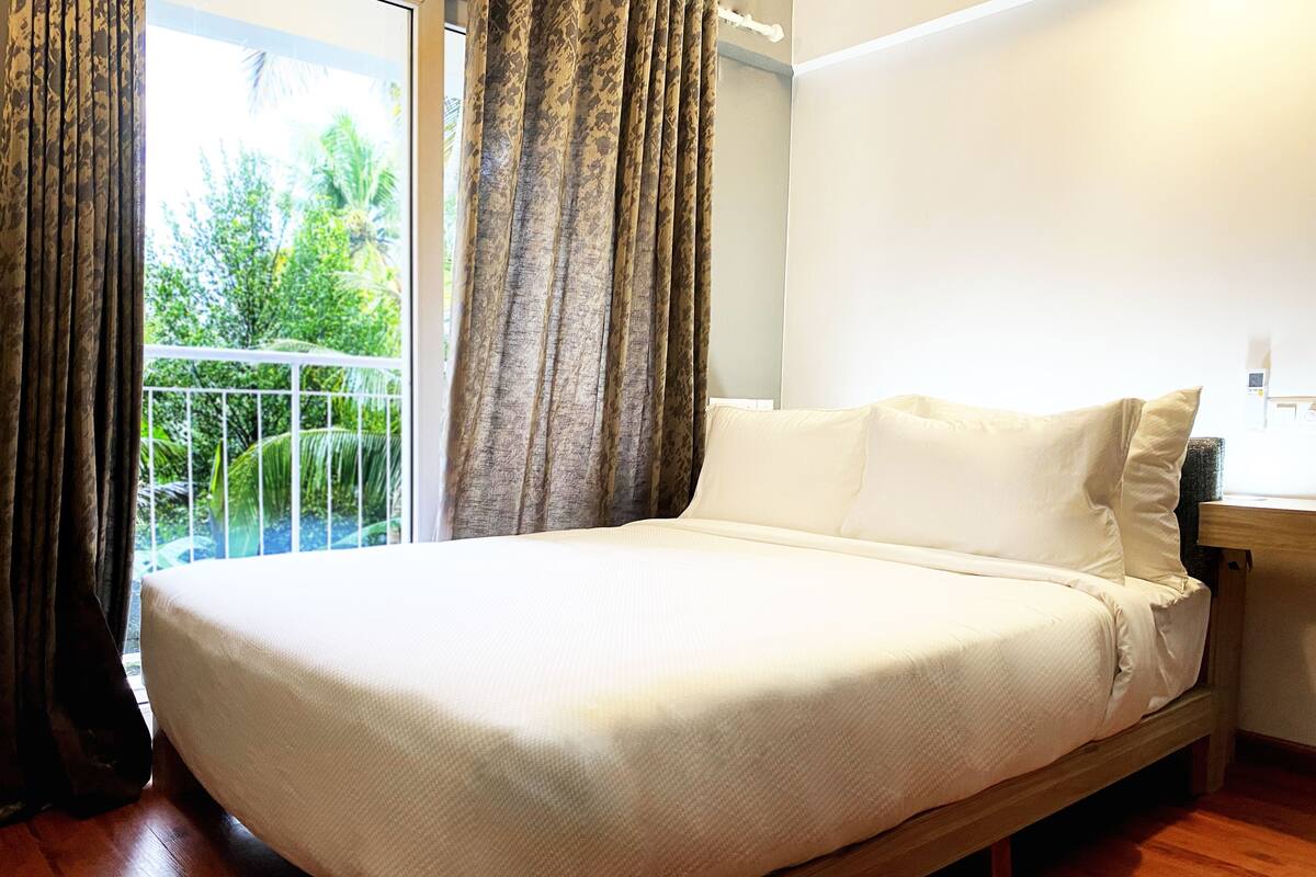 A bed with a white mattress and pillows, a wooden headboard, and a view of greenery through a window with curtains.