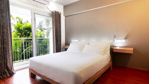 Una cama bien tendida en una habitación con suelo de madera, un balcón con cortinas y una lámpara fijada a la pared.