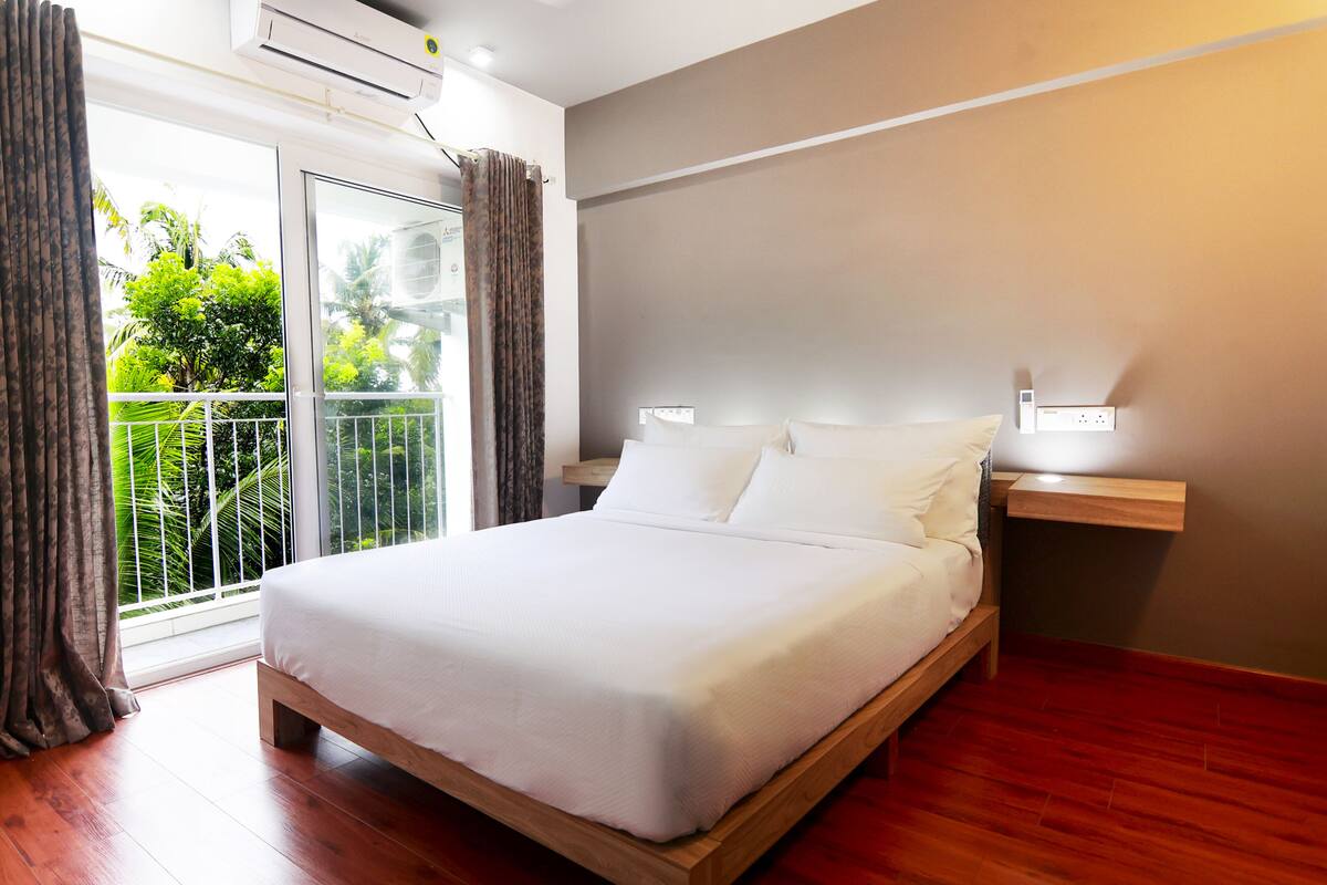 A neatly made bed in a room with wooden flooring, a balcony with curtains, and a wall-mounted light fixture.