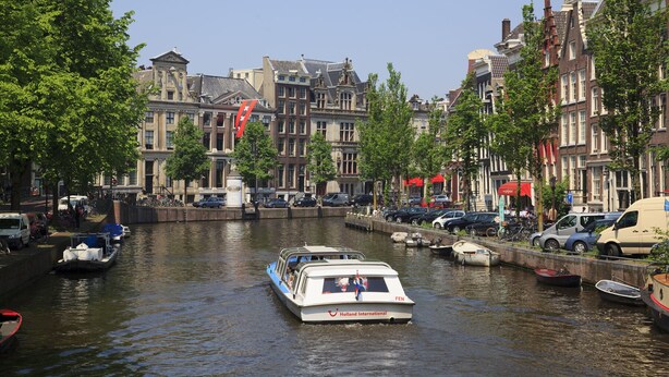 Un canal con una embarcación llamada 'Holland International' en primer plano, rodeado de edificios históricos y coches estacionados a lo largo de la orilla.