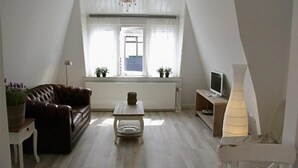 Una sala de estar pequeña y bien iluminada, con techo inclinado, un sofá, una mesita de centro, una lámpara y un televisor.