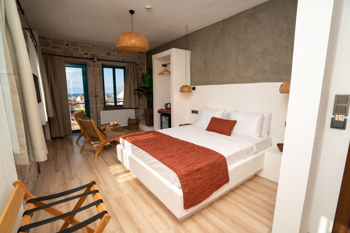 A bedroom with a large bed, a wooden chair, a hanging light fixture, and a view of the outside through a window.