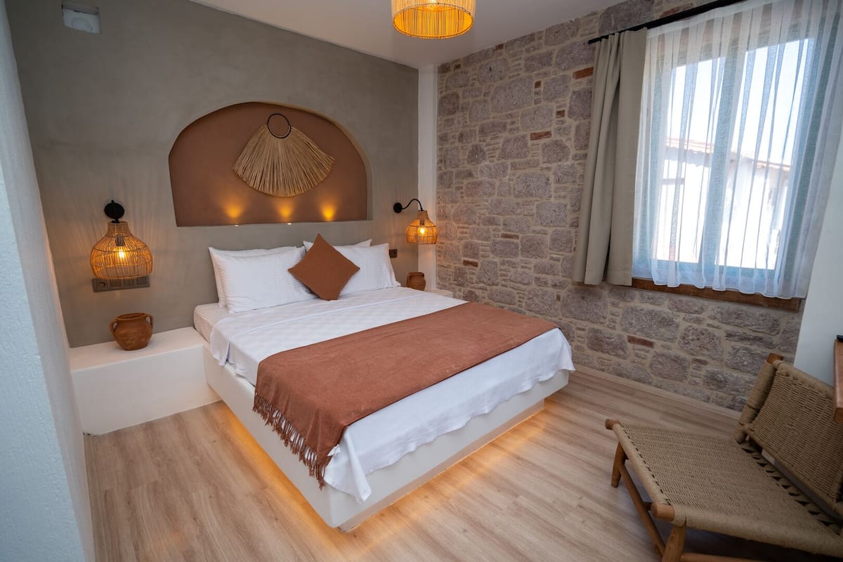 A bedroom with a stone wall, a bed with white bedding and a brown throw, two wall-mounted lamps, and a wicker chair.