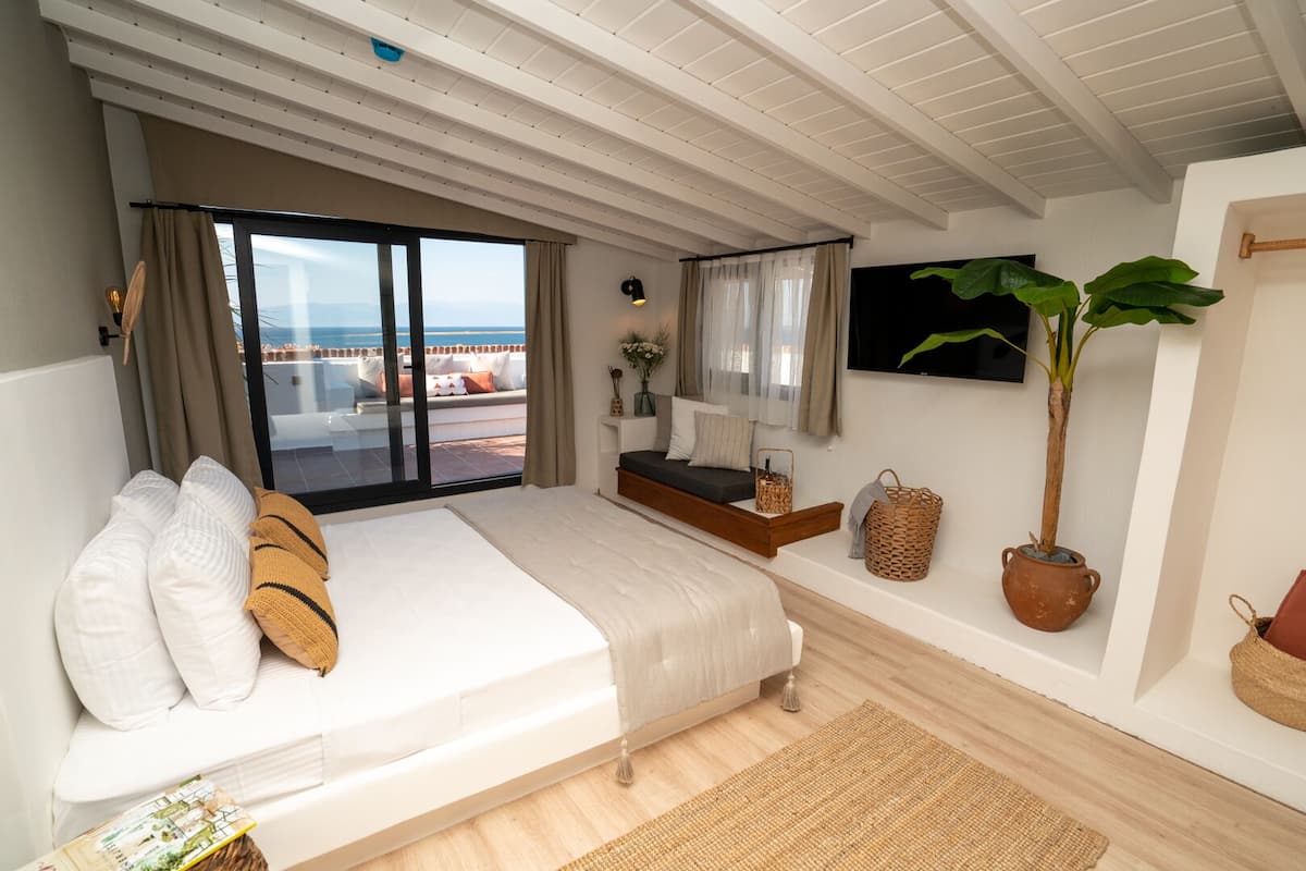 A bedroom with a large bed, a small bedside table, a potted plant, and a view of the sea.