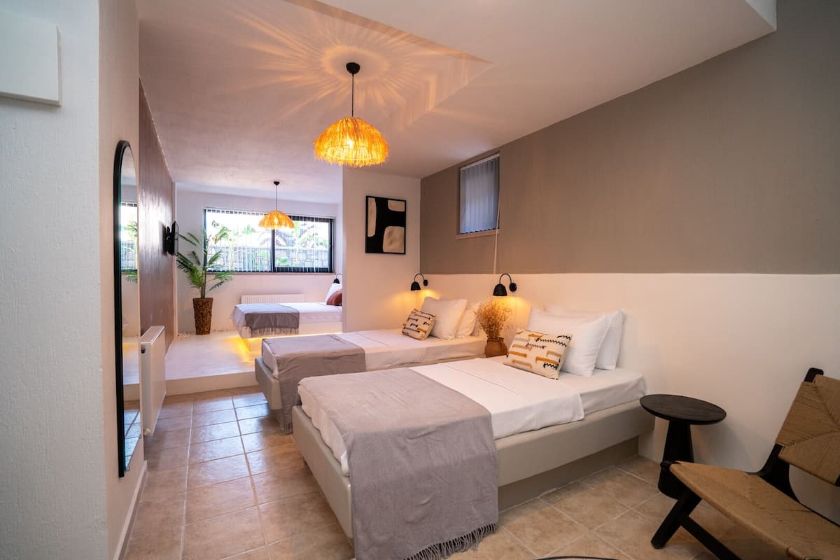A modern bedroom with two beds, a small table, a chair, and a large window with a view of greenery.